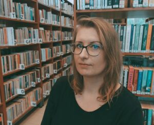 Olga blondynka stoi na tle regałów bibliotecznych